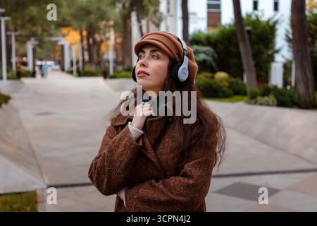 Moment de loisir urbain avec une femme caucasienne élégante marchant calmement et appréciant la musique à travers ses écouteurs dans les rues tranquilles de la ville d'automne Banque D'Images