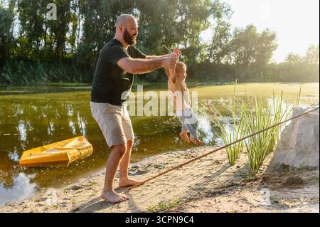 Homme souriant jouant avec un petit enfant au bord de la rivière au coucher du soleil. Père tient l'enfant en bas âge en l'air tout en se tenant pieds nus sur la rive sablonneuse avec vert Banque D'Images
