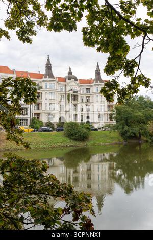 Élégant manoir historique avec des tours ornées et des toits de tuiles rouges reflétés dans l'eau calme de la rivière encadrée par des branches d'arbres vertes. Européenne architectural Her Banque D'Images
