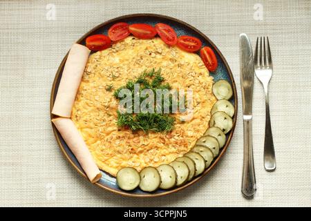 Une omelette à l'avoine repose sur une assiette bleue à côté de concombres légèrement salés, de tomates et de jambon. Vue de dessus. Gros plan. Banque D'Images