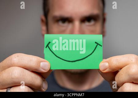 Allemagne - 27 septembre 2025 : un homme tient un Post-it vert avec un visage souriant, symbole de bonheur, de joie et d'optimisme. *** Mann hält ein Grünes Post-it mit einem lächelnden Smiley vor sich Symbol für Glück, Freude und Optimismus. Banque D'Images