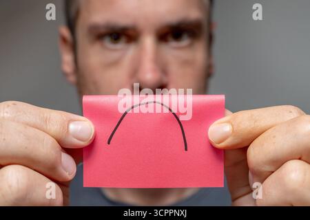 Allemagne - 27 septembre 2025 : un homme tient un morceau de papier avec un triste symbole dessus. L'image symbolise la dépression, l'épuisement professionnel et le stress mental qui affectent le psychisme et la vie quotidienne. *** Ein Mann hält ein papier mit einem traurigen symbole vor sich. DAS Bild steht sinnbildlich für Depression, Burnout und mentale Belastungen, die auf Psyche und Alltag wirken. Banque D'Images