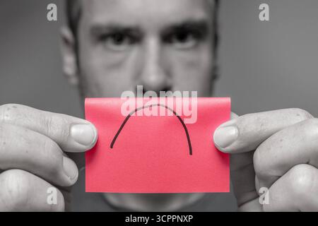 Allemagne - 27 septembre 2025 : un homme tient un morceau de papier avec un symbole triste à la caméra. L'image symbolise le mécontentement politique en Allemagne et l'humeur dépressive dans la société. *** Ein Mann hält ein Zettelchen mit traurigem symbole in die Kamera. DAS Bild steht sinnbildlich für politische Unzufriedenheit in Deutschaland und die gedrückte Stimmung in der Gesellschaft. Banque D'Images