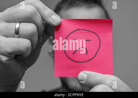 Allemagne - 27 septembre 2025 : un homme tient un morceau de papier avec un triste symbole dessus. L'image symbolise l'humeur dépressive de nombreuses personnes en raison des crises actuelles telles que la pandémie, la guerre en Ukraine, la menace de guerre et l'inflation en Allemagne. Il représente l'insatisfaction dans la vie quotidienne ou au travail et illustre la frustration, la surcharge et un manque de contentement. *** Ein Mann hält ein Zettelchen mit traurigem symbole vor sich. DAS Bild symbolisiert die gedrückte Stimmung vieler Menschen durch aktuelle Krisen wie Pandemie, Krieg in der Urkaine und Kriegsgefahr und inflation in Deutschla Banque D'Images