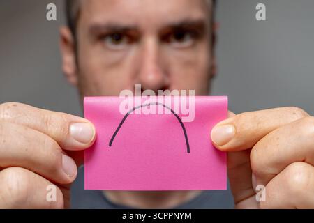 Allemagne - 27 septembre 2025 : un homme tient un morceau de papier avec un triste symbole dessus. L'image symbolise l'humeur dépressive de nombreuses personnes en raison des crises actuelles telles que la pandémie, la guerre en Ukraine, la menace de guerre et l'inflation en Allemagne. Il représente l'insatisfaction dans la vie quotidienne ou au travail et illustre la frustration, la surcharge et un manque de contentement. *** Ein Mann hält ein Zettelchen mit traurigem symbole vor sich. DAS Bild symbolisiert die gedrückte Stimmung vieler Menschen durch aktuelle Krisen wie Pandemie, Krieg in der Urkaine und Kriegsgefahr und inflation in Deutschla Banque D'Images