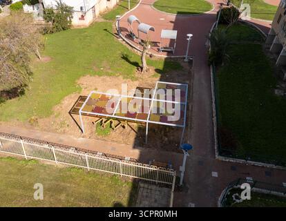 Vue aérienne d'un abri coloré de terrain de jeu projetant des ombres sur l'herbe, contrastant avec les motifs géométriques des allées environnantes, Nahariyya, District Nord, Israël. Banque D'Images