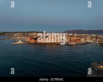 Vue aérienne du port vénitien projetant des teintes chaudes sur l'eau, avec le phare debout contre le ciel crépusculaire, Chania, Chania, Grèce. Banque D'Images