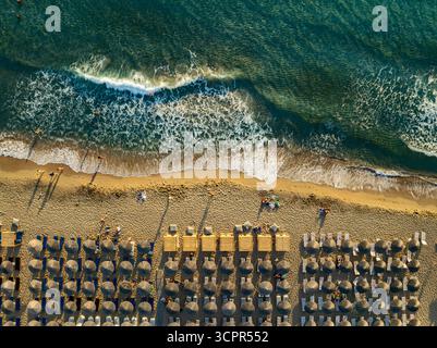 Vue aérienne des parasols baignés de soleil bordent le sable doré où les vagues mousseuses s'écrasent à terre, peignant une scène pittoresque de bonheur balnéaire, Chania, Chania, Grèce. Banque D'Images
