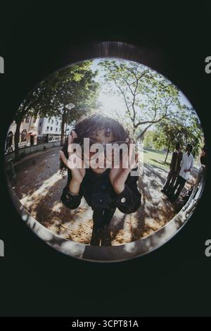 Fish Eye Lens portrait de jeune femme à la mode portant des lunettes de soleil tout en se tenant dans le parc à la journée ensoleillée Banque D'Images