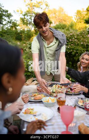 Heureux jeune homme gardant le bol de salade de pâtes sur la table au dîner avec des amis dans la cour arrière Banque D'Images
