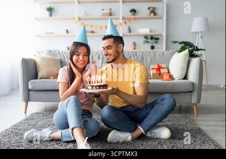 Joyeux couple millénaire portant des chapeaux de fête, célébrant l'anniversaire avec un gâteau de fête à la maison, sur toute la longueur Banque D'Images