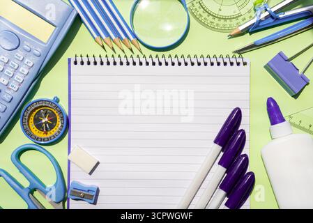 Image horizontale d'un grand cahier entouré de stylos violets, ciseaux, colle, règle, boussole, et taille-crayon accentué violet sur fond vert. Parfait pour l'école, le bureau, les projets créatifs ou la planification d'études. Banque D'Images