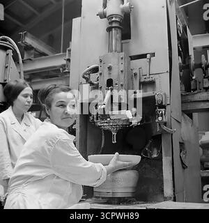 Deux travailleuses en manteau blanc exploitent une grande presse industrielle pour former des isolateurs en céramique à l'usine de raccords et isolateurs de Slavyansk (AIZ) en RSS d'Ukraine dans les années 1970 Cette photo d'archive monochrome est un portrait d'une équipe ouvrière soviétique qualifiée, avec les femmes gérant avec confiance les machines complexes. La scène présente la production en série de composants essentiels pour le réseau d'électrification de l'URSS et met en évidence le rôle vital des femmes dans l'industrie lourde de la région du Donbass Banque D'Images