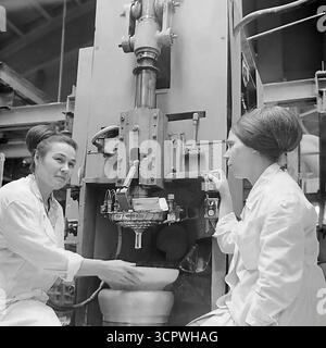 Deux travailleuses en manteau blanc exploitent une grande presse industrielle pour former des isolateurs en céramique à l'usine de raccords et isolateurs de Slavyansk (AIZ) en RSS d'Ukraine dans les années 1970 Cette photo d'archive monochrome est un portrait d'une équipe ouvrière soviétique qualifiée, avec les femmes gérant avec confiance les machines complexes. La scène présente la production en série de composants essentiels pour le réseau d'électrification de l'URSS et met en évidence le rôle vital des femmes dans l'industrie lourde de la région du Donbass Banque D'Images