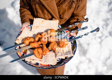 Personne tenant une assiette avec de la viande grillée brochée et du pain plat à l'extérieur dans la neige. Banque D'Images
