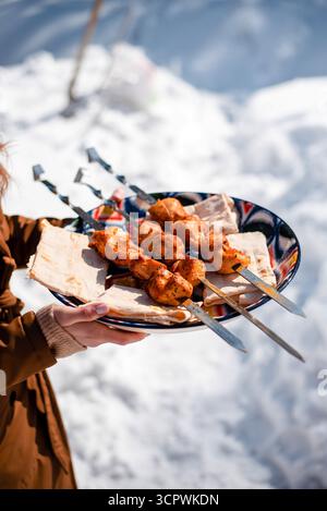Personne tenant une assiette avec des brochettes grillées et du pain plat dans un cadre extérieur enneigé. Banque D'Images