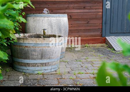 À côté d'une maison, une rampe en métal s'élève de carreaux de pavage tandis que deux vieux barils en bois avec des bandes métalliques tiennent de petites fontaines à tuyaux. De l'eau claire tombe de th Banque D'Images