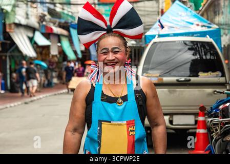 Bangkok, Thaïlande, 5 septembre 2025 : vendeur de rue thaïlandais joyeux avec un maquillage audacieux et un arc géant aux couleurs du drapeau thaïlandais. Banque D'Images
