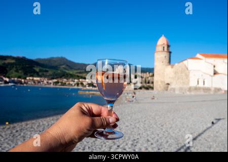 Main avec verre de vin rose Collioure AOC aux rayons du soleil avec vue sur la vieille ville de Collioure, maisons colorées, église, château et plages, Occitanie, franc Banque D'Images