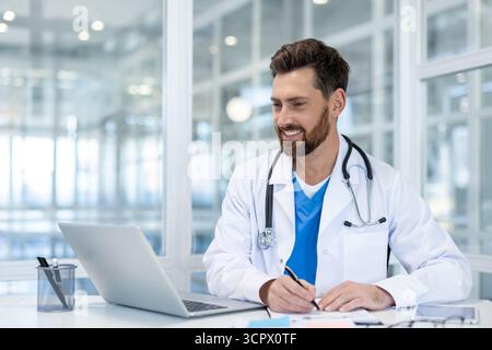 Médecin masculin confiant en manteau blanc au bureau souriant à l'ordinateur portable pendant le rendez-vous de télémédecine tout en prenant des notes, fournissant des consultations médicales à distance et des soins (télésanté) Banque D'Images