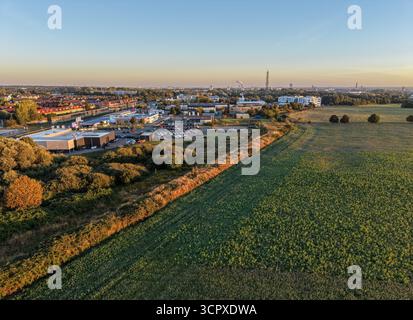 Vue aérienne de la périphérie d’une ville où l’industrie rencontre des champs verts ouverts, baignés de lumière chaude du matin avec une ligne d’horizon lointaine sous un ciel clair Banque D'Images