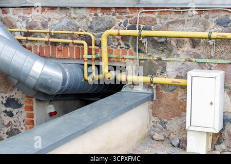 Gros plan d'un grand conduit argenté et de tuyaux utilitaires jaune vif montés sur un mur rustique de pierre et de brique mélangées. Banque D'Images