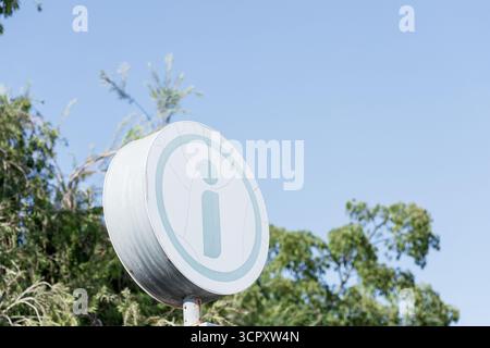 Un panneau d'information circulaire portant le symbole « i » bleu clair est monté sur un poteau, placé contre un ciel bleu clair et un feuillage vert flou. Banque D'Images