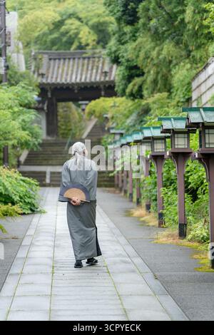 Un vieil homme dans un kimono tenant un ventilateur pliant marche sur un chemin de pierre vers une porte de temple. Kyoto, Japon. Banque D'Images