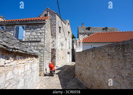 Un scooter rouge vif repose le long d'une rue pavée ensoleillée encadrée par des murs de pierre dans un village historique. textures rustiques, ruelles étroites et lumière chaude e Banque D'Images