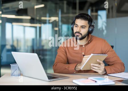 Jeune homme portant des écouteurs et utilisant un ordinateur portable pour l'apprentissage en ligne ou un webinaire, se concentrant intentionnellement sur une conférence virtuelle et notant avec diligence des notes importantes dans un bureau moderne Banque D'Images