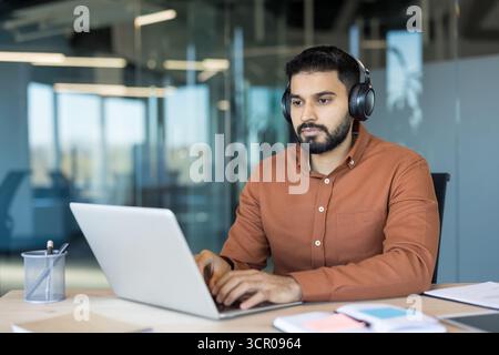 Jeune homme d'affaires indien portant des écouteurs, se concentrant intentionnellement tout en tapant sur un ordinateur portable, travaillant à un bureau propre dans un environnement de bureau contemporain ouvert Banque D'Images