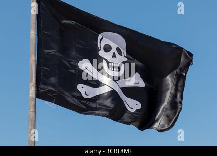 Drapeau noir avec crâne blanc et os croisés ondulant au vent contre un ciel bleu clair. La surface brillante suggère une matière plastique. Banque D'Images