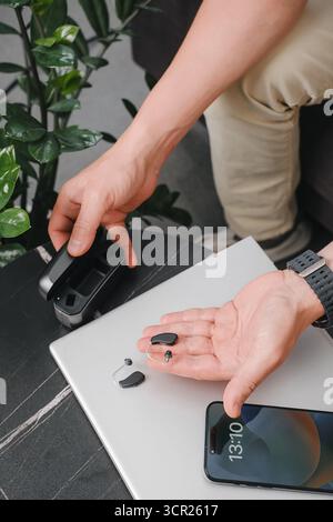 Aides auditives numériques modernes, smartphone et étui de chargement sur une table de bureau élégante, avec une personne préparant les appareils pour le travail. Environnement professionnel Banque D'Images