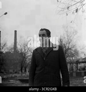 Un portrait puissant et poignant en plein air d'un travailleur âgé et altéré par les intempéries, son visage profondément bordé racontant l'histoire d'une longue vie de travaux forcés, posant sur fond d'une grande usine avec des cheminées à Sloviansk, RSS d'Ukraine, vers 1985. Cette profonde photographie en noir et blanc capture la dignité tranquille et la résilience d'un vétéran de l'industrie soviétique, personnifiant la génération qui a construit la puissance industrielle de la région et servant d'hommage au fondement humain du Donbass pacifique Banque D'Images