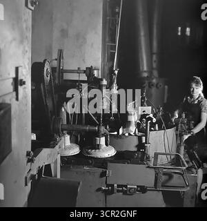 Une jeune travailleuse, souriante avec fierté et concentration, exploite une ligne de production semi-automatique d'isolateurs en céramique dans un atelier éclairé de l'usine d'armature et d'isolateur de Slavyansk (AIZ), vers 1985. Cette photographie atmosphérique en noir et blanc de Sloviansk, RSS d'Ukraine, capture magnifiquement la synergie entre l'homme et la machine dans l'industrie soviétique, soulignant le rôle essentiel de la main-d'œuvre féminine qualifiée et symbolisant l'esprit intentionnel et créatif du Donbass pacifique. Banque D'Images