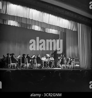 Un orchestre symphonique, dirigé par un chef d'orchestre et mettant en vedette une femme soliste, donne un concert de musique classique sur la scène d'un Palais de la culture à Sloviansk, RSS d'Ukraine, vers 1985. Cette photographie d'ambiance en noir et blanc, prise du point de vue du public, capture un événement culturel important dans une ville industrielle soviétique, avec un slogan patriotique visible au-dessus de la scène, symbolisant la promotion de l'art et la riche vie culturelle de la paisible région du Donbass Banque D'Images