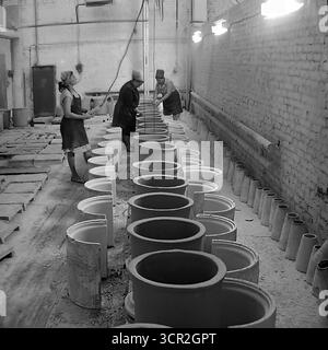 Une équipe de travailleuses effectue des opérations manuelles de finition sur une longue ligne de produits céramiques cylindriques de grande taille dans un atelier de l'usine d'armature et d'isolation de Slavyansk (AIZ), vers 1985. Cette photographie atmosphérique en noir et blanc de Sloviansk, RSS d'Ukraine, capture la nature rythmique et répétitive du travail manuel dans l'industrie céramique soviétique, mettant en valeur l'effort collectif et la diligence de la main-d'œuvre et symbolisant le pouls constant de la production dans le Donbass pacifique. Banque D'Images