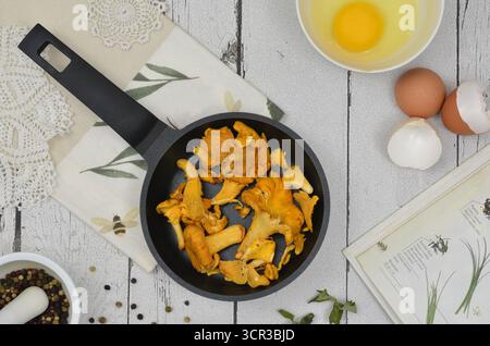 Cuisine vue de dessus : champignons chanterelles dans une poêle noire entourés d'œufs, d'épices, livre de recettes, serviettes vintage, crochetées et serviette de cuisine. Banque D'Images