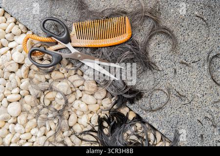 Le coiffeur. Ciseaux avec peigne brun et boucle de cheveux gris sur un fond de roche noire. Vue de dessus. Banque D'Images