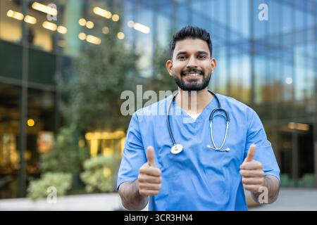 Jeune médecin indien souriant portant des gommages bleus et un stéthoscope, debout à l'extérieur devant un bâtiment moderne, montrant un signe pouce vers le haut positif avec les deux mains Banque D'Images