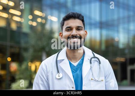 Jeune médecin indien portant une blouse de laboratoire blanche et un stéthoscope, souriant avec confiance à la caméra en plein air avec un bâtiment hospitalier moderne en arrière-plan Banque D'Images