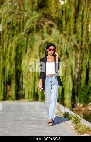 Femme en tenue décontractée marchant dehors, portant des lunettes de soleil rouges et utilisant un smartphone. Espagne Banque D'Images