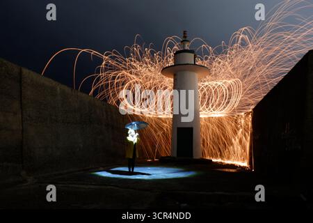 Peinture légère en laine d'acier avec parapluie rose – étincelles d'art par le phare. Une femme tenant un parapluie rose se tient devant un phare AS Banque D'Images