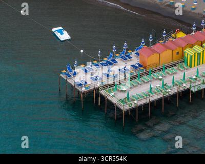 Vue aérienne des parasols de plage vibrants et des chaises longues bordent des piliers en bois s'étendant dans la mer turquoise, contrastant avec les cabanes de plage colorées, Sorrente, Campanie, Italie. Banque D'Images