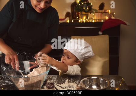 Une mère heureuse guide son enfant alors qu'ils mélangent la farine et la pâte pour une maison en pain d'épices, portant un chapeau de chef dans une cuisine festive et chaleureusement éclairée. Noël de Banque D'Images