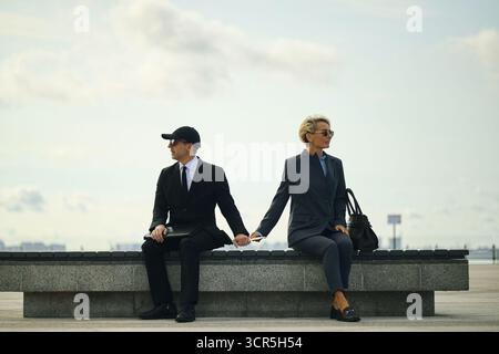 Jeune homme d'affaires caucasien et femme mature assis sur le banc tout en faisant face à l'autre, en passant lettre ou document, à la fois portant des vêtements d'affaires et des lunettes de soleil à l'extérieur Banque D'Images