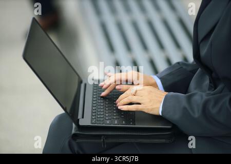 Femme d'affaires d'âge moyen en costume formel tapant sur ordinateur portable à l'extérieur, mains reposant sur le clavier, portant des vêtements d'affaires, assis sur le banc, se concentrant sur le travail, cadre professionnel Banque D'Images