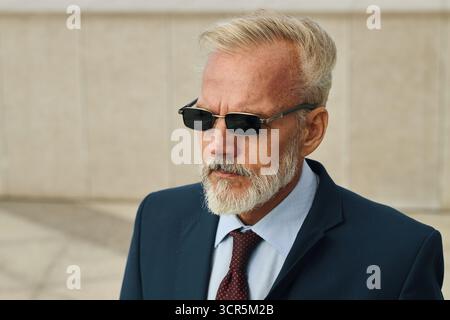 Homme senior caucasien portant des lunettes de soleil marchant à l'extérieur, portant les cheveux gris courts et la barbe, habillé en costume formel et cravate, maintenant l'expression confiante sérieuse, sur le chemin des négociations d'affaires Banque D'Images