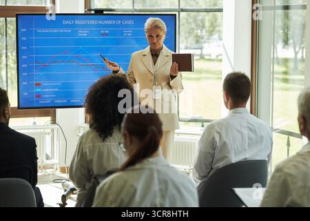 Cadre de clinique médicale féminine caucasienne présentant des données sur grand écran à divers groupes de jeunes adultes et professionnels d'âge moyen, tenant une tablette numérique et faisant des gestes vers l'affichage graphique Banque D'Images