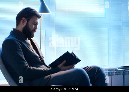 Architecte à la recherche de nouvelles idées. Homme barbu élégant confiant en costume et col roulé assis dans une chaise souple dans le bureau et la lecture du magazine sur les nouvelles tendances dans la conception moderne des bâtiments Banque D'Images